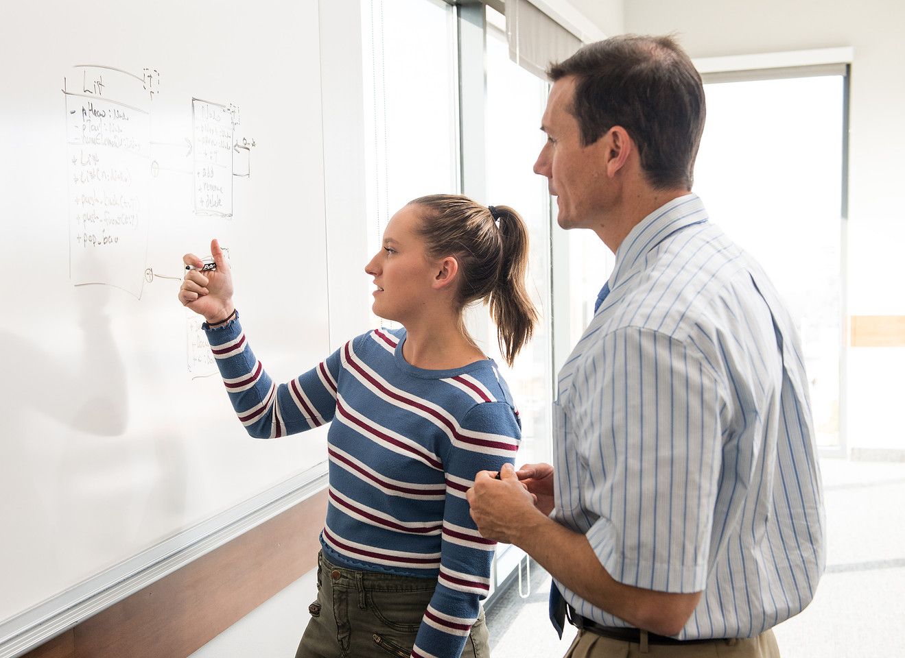 A student explains an idea at a whiteboard while an instructor listens